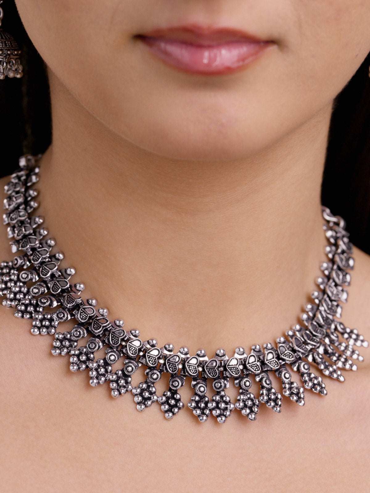 Close-up of a model wearing a minimal oxidised necklace with raised heart-shaped upper links, textured metalwork, and a patterned lower fringe sitting close to the neckline.