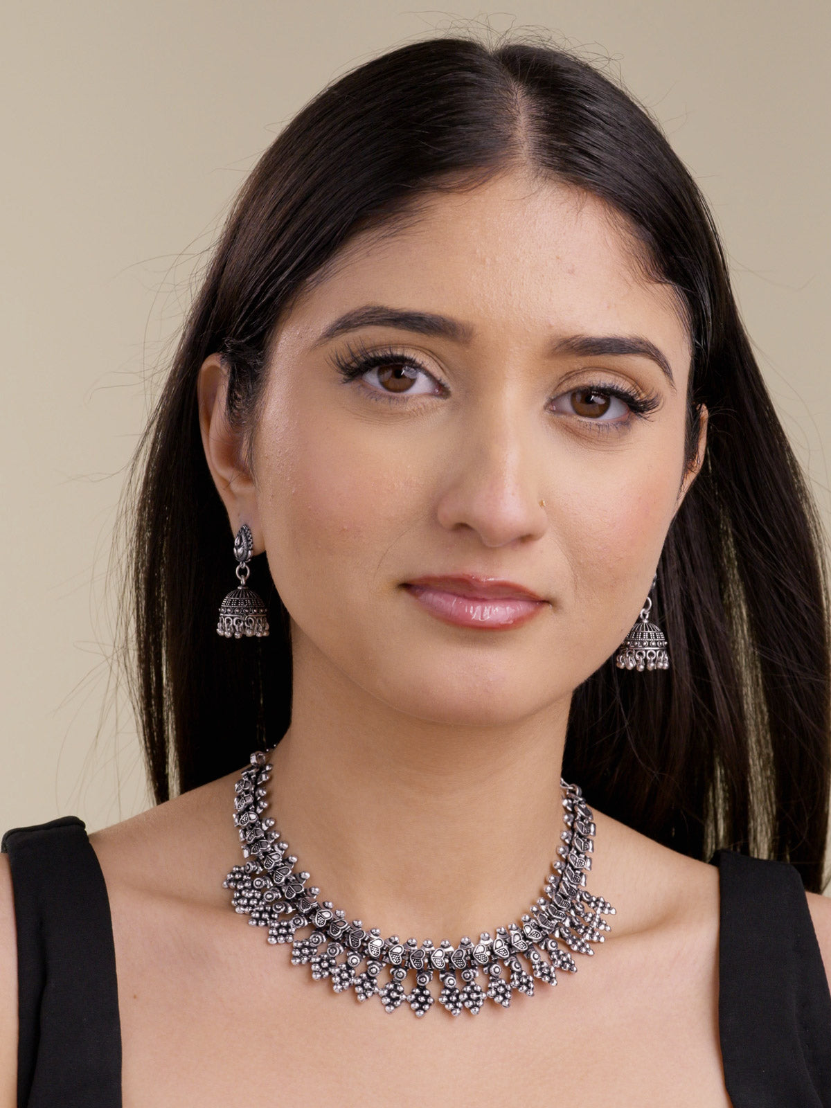 Model wearing a minimal oxidised necklace set with matching small jhumki earrings, raised heart-shaped upper links, textured metalwork, and a patterned lower fringe in a front portrait shot.