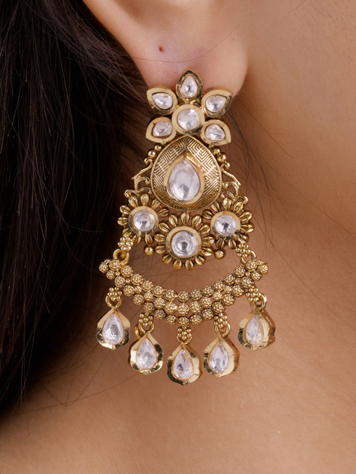 Extreme close-up of Rajwadi earring on a model ear with kundan-look stones, textured centre teardrop detail, floral round accents, and layered pear-shaped dangles in matte gold tone.