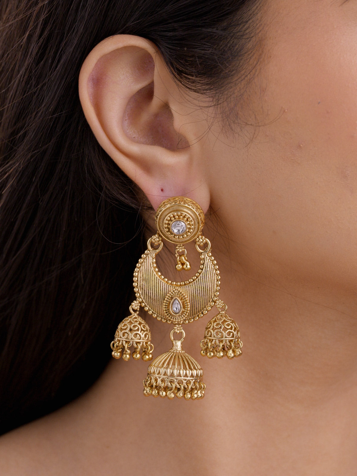 Close-up of Rajwadi earring on a model ear with matte gold tone detailing, white kundan-look stones, ribbed crescent centre panel, and three hanging jhumki drops.