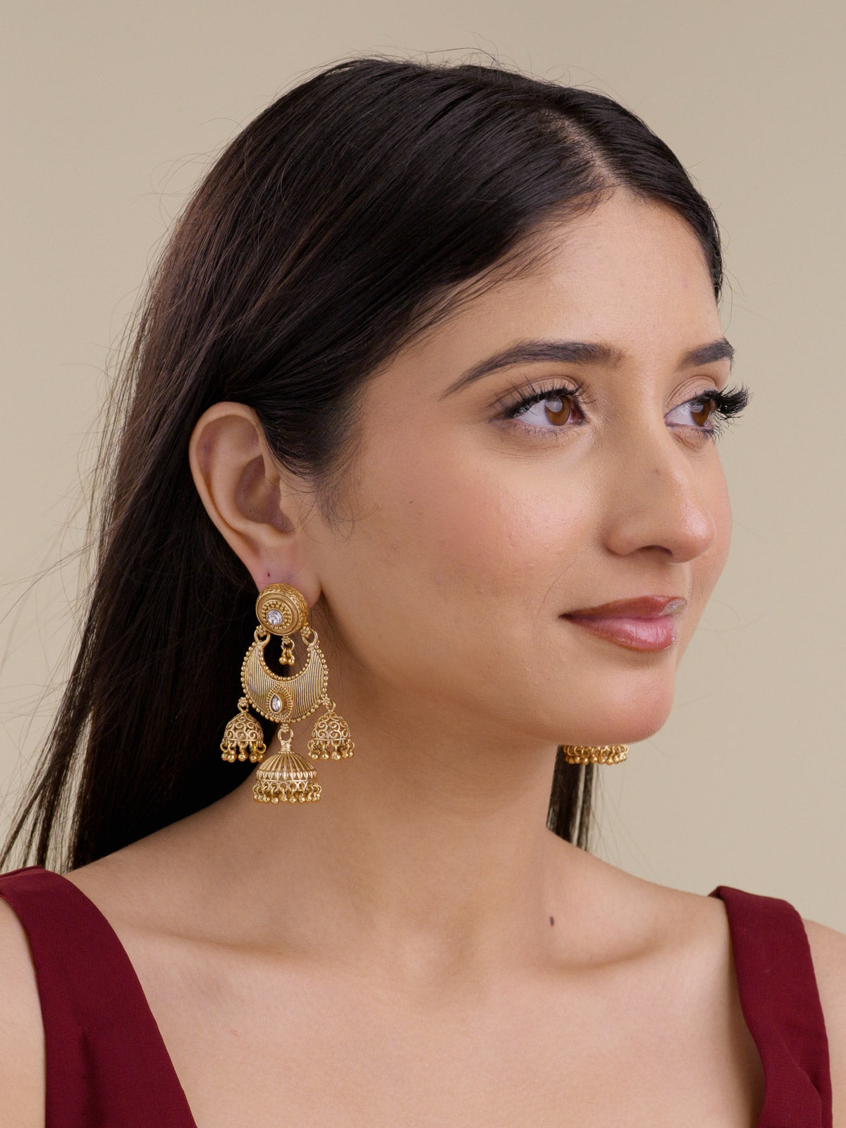 Model wearing Rajwadi earrings in matte gold tone with white kundan-look stones, crescent centre detailing, and multi-jhumki drops styled with a burgundy outfit.