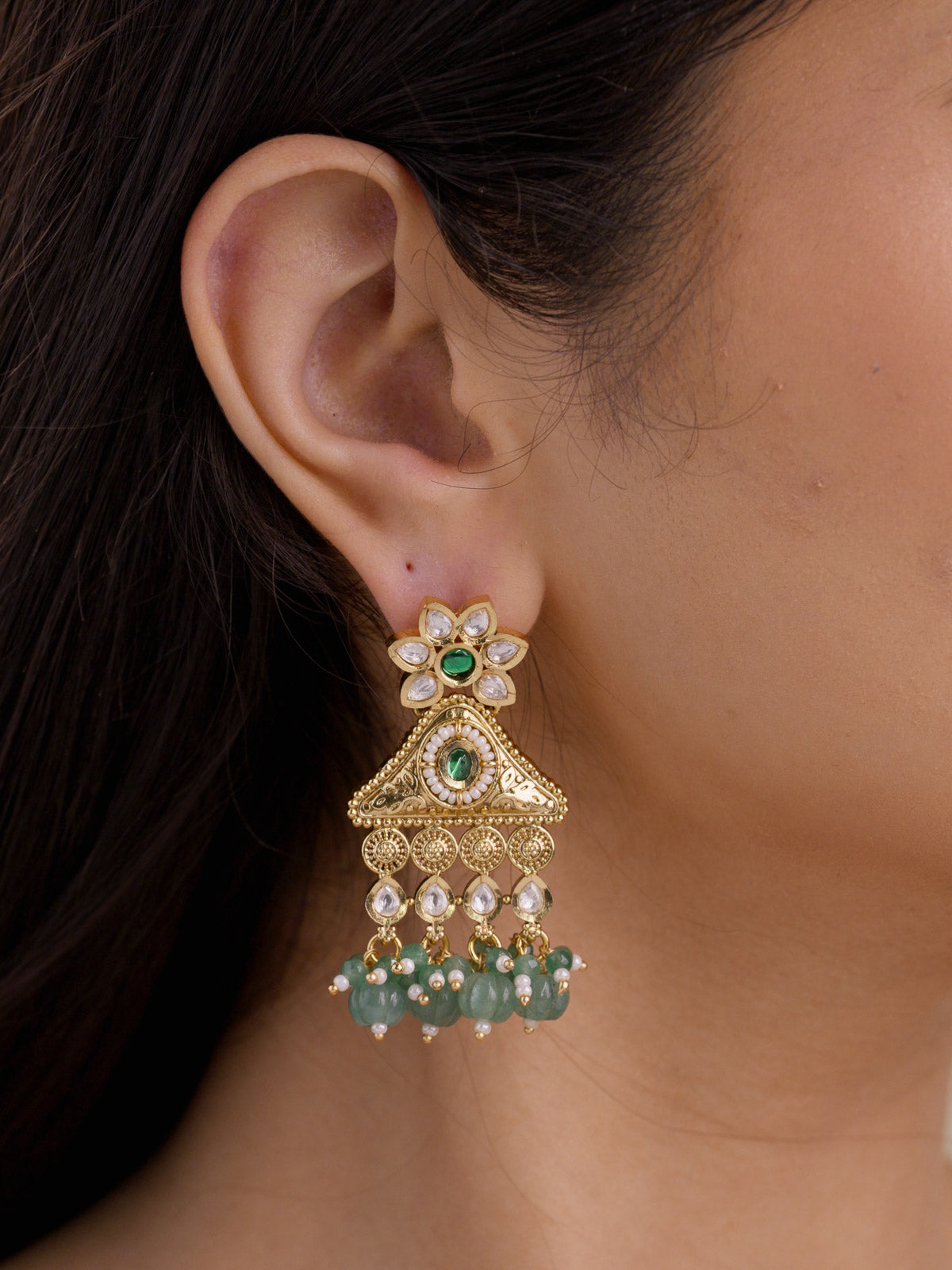 Close-up side view of Rajwadi earring on a model ear with kundan-look stones, green centre accents, triangular matte gold tone detailing, pearl-style accents, and sage green rounded drops.
