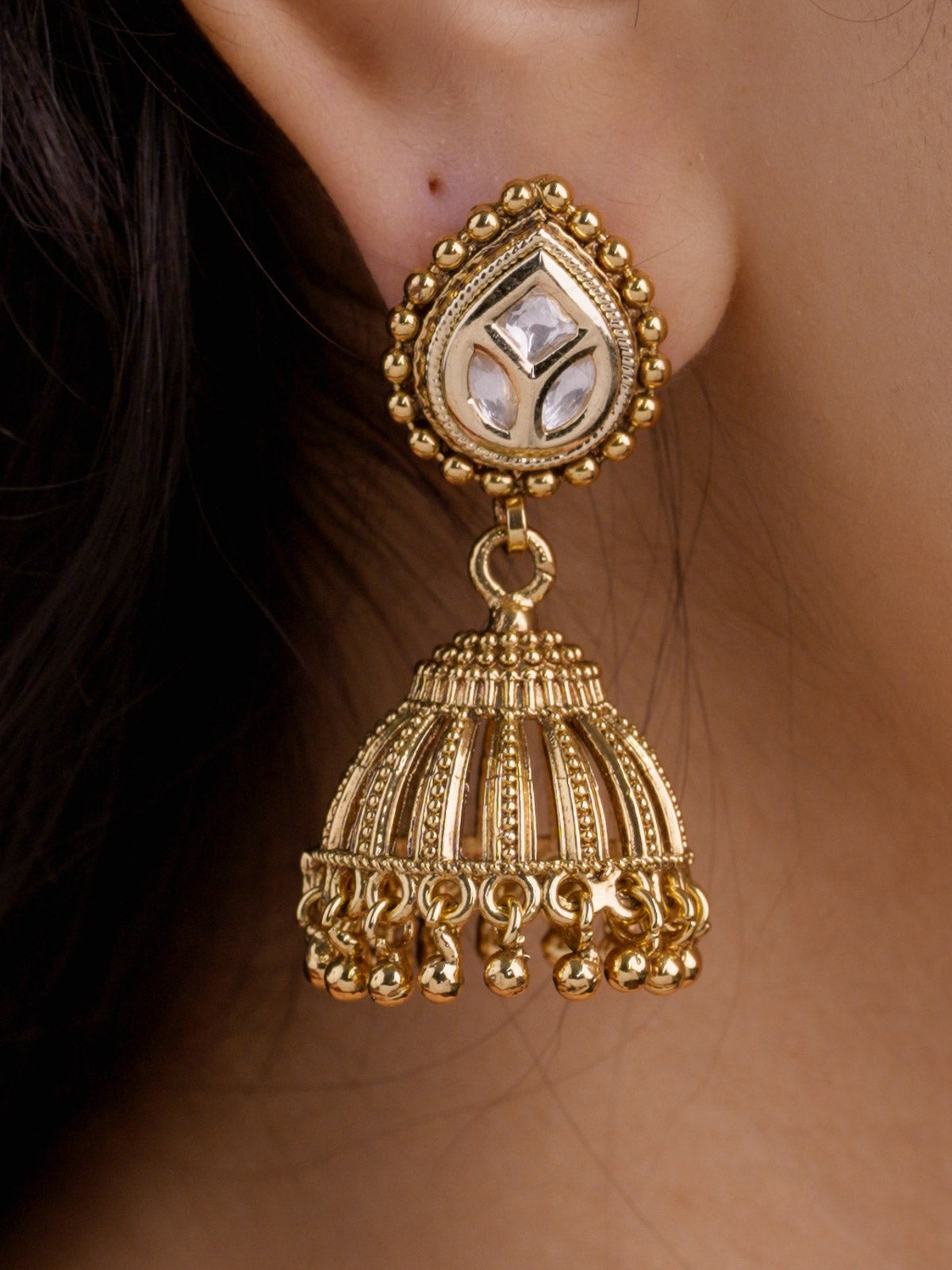 Close-up of matte gold Rajwadi earring on a model ear with kundan-look stones in the teardrop stud, raised gold edging, domed jhumka lower section, and small gold ball dangles.