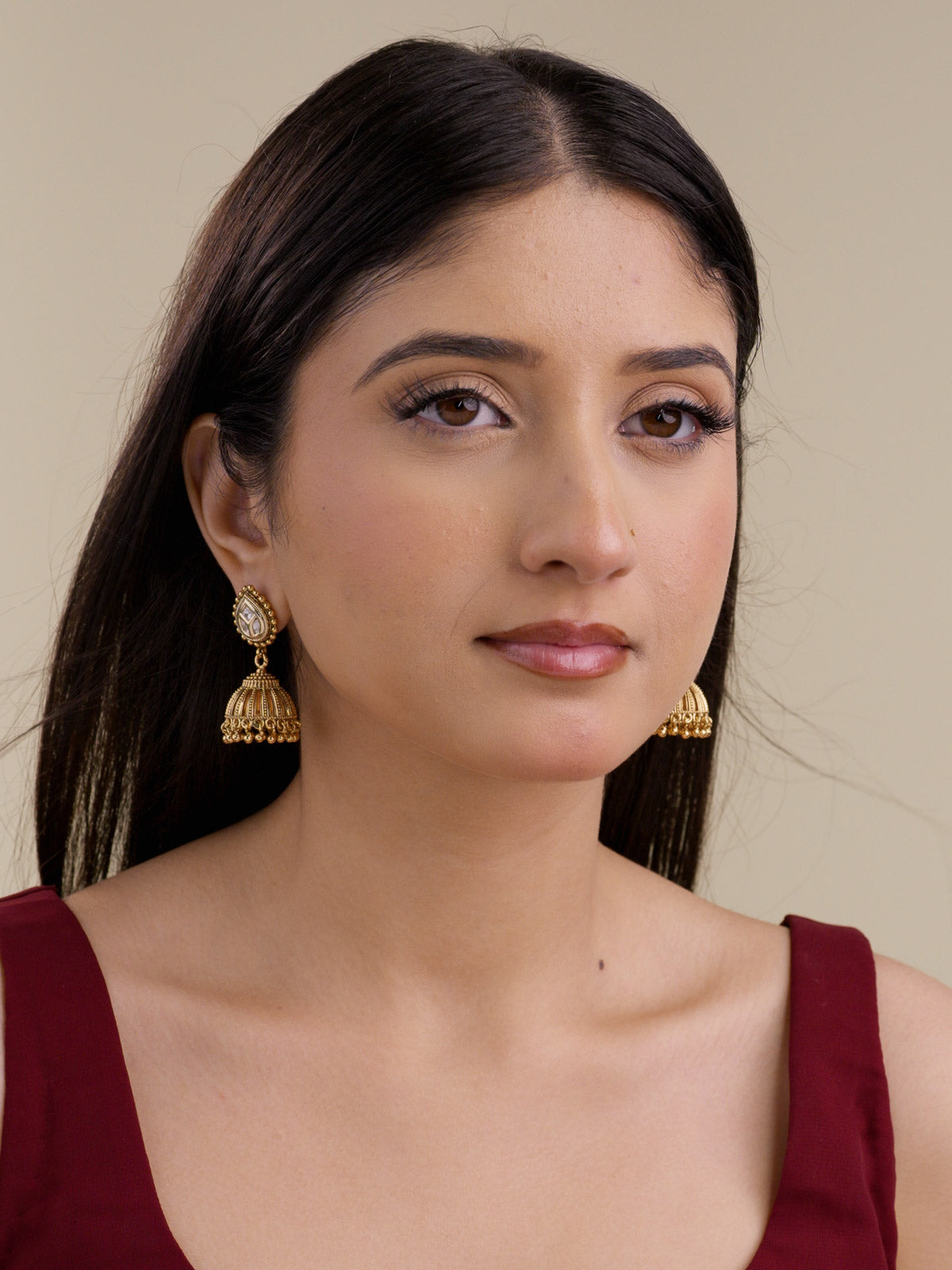 Front view of model wearing matte gold Rajwadi jhumka earrings with teardrop stud tops, kundan-look stones, domed lower sections, and small gold ball dangles styled with a burgundy outfit.