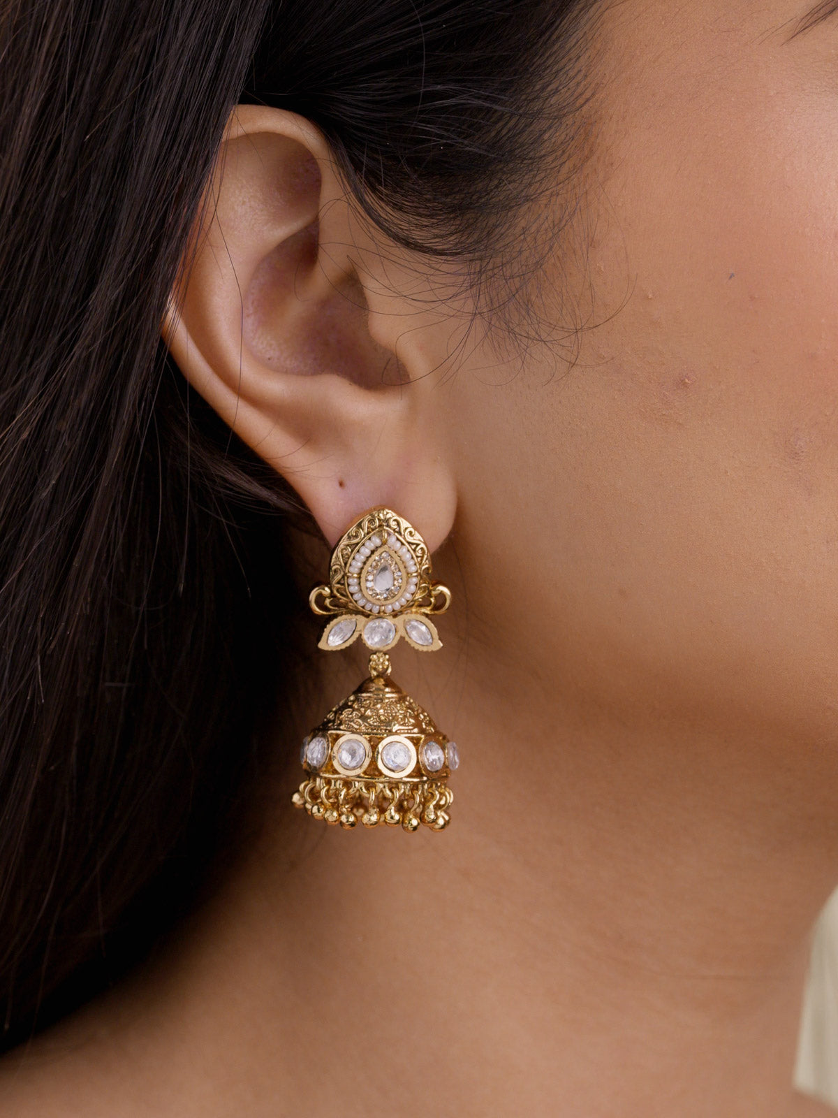 Model ear wearing a gold tone Rajwadi jhumka earring with teardrop kundan-look detailing, pearl-style border, leaf-shaped clear stones, textured dome, and small gold ball fringe.
