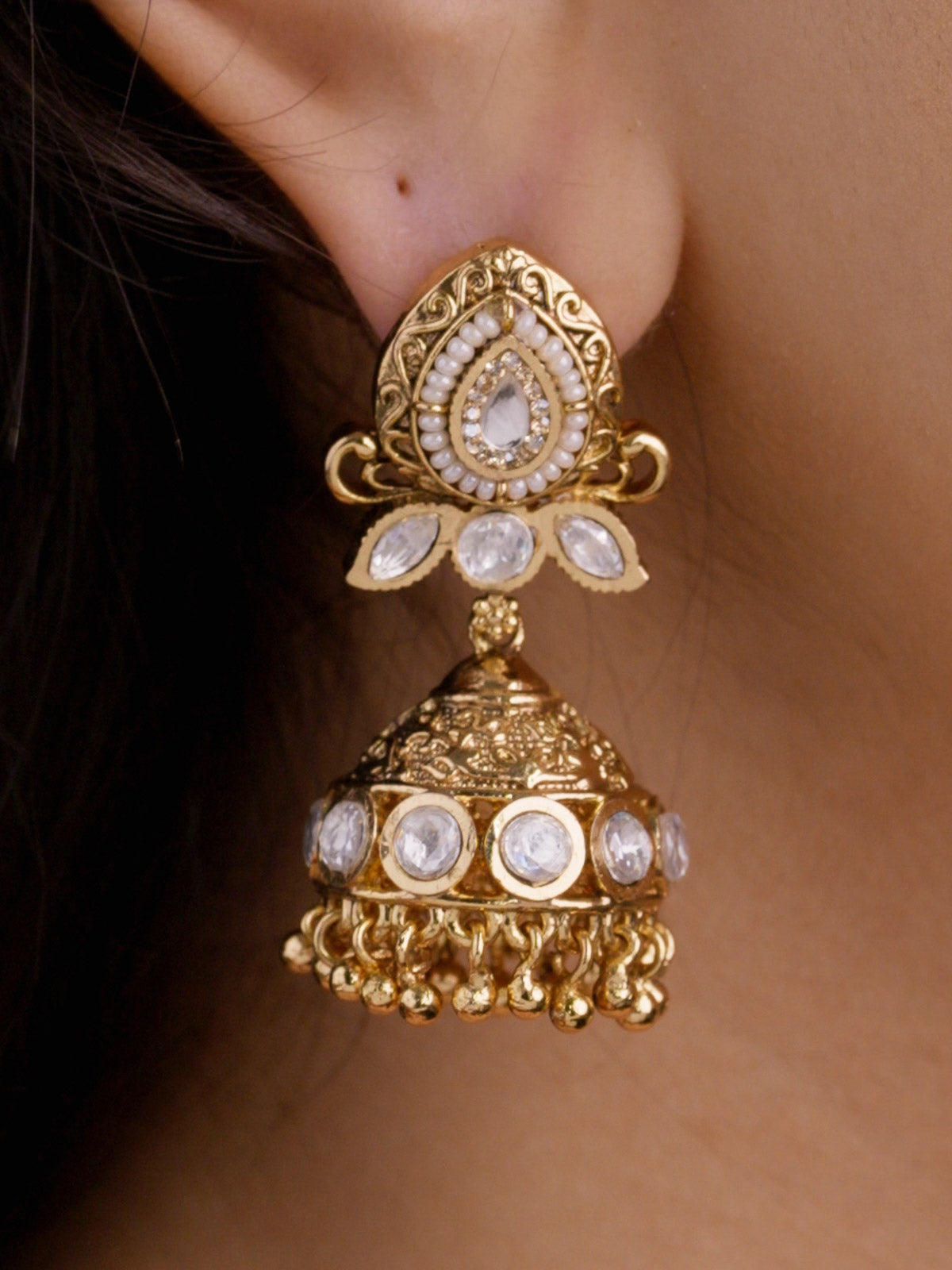 Close-up of Rajwadi jhumka earring on a model ear with teardrop kundan-look centre, pearl-style beaded border, leaf-shaped clear stones, dome detail, and gold ball fringe in gold tone.