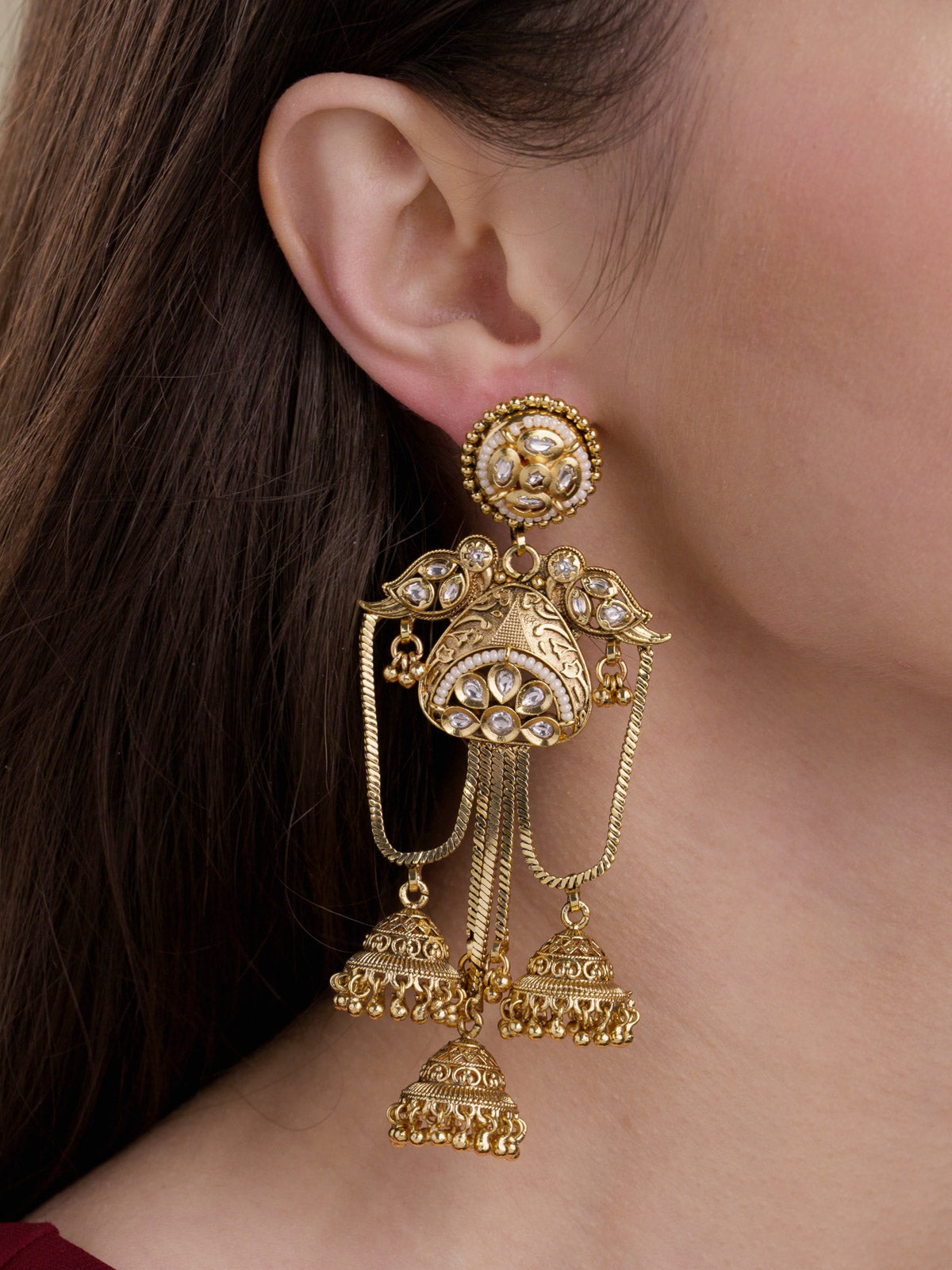 Close-up of a model wearing matte gold tone Rajwadi earrings with round stud tops, clear stone detailing, bird-inspired side motifs, chain drapes and three bell drops.
