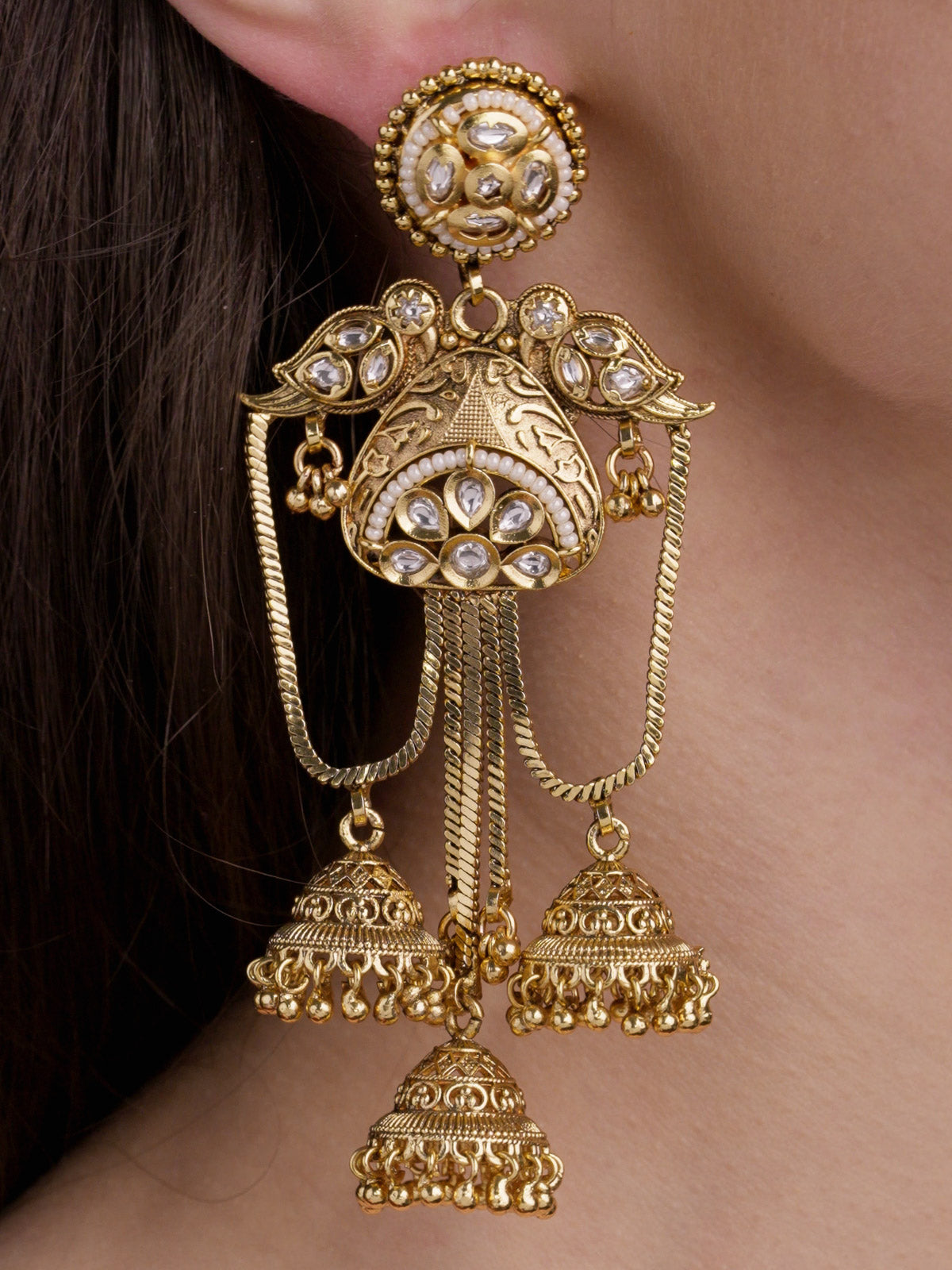 Close-up of a model wearing a matte gold tone Rajwadi earring with a round stud top, clear stone detailing, bird-inspired side motifs, chain drapes and three bell-shaped jhumka drops.