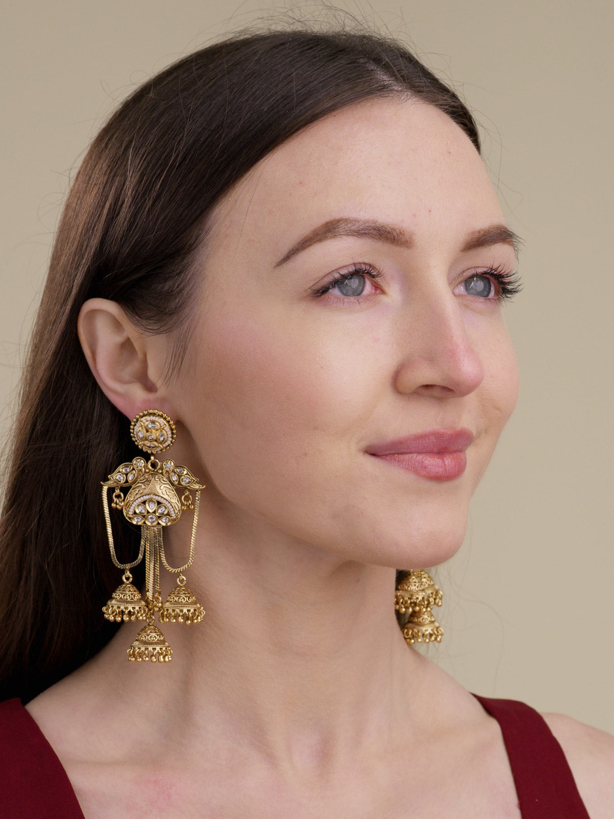 Model wearing matte gold tone Rajwadi statement earrings with round stud tops, clear stone detailing, bird-inspired side motifs, chain drapes and bell drops.