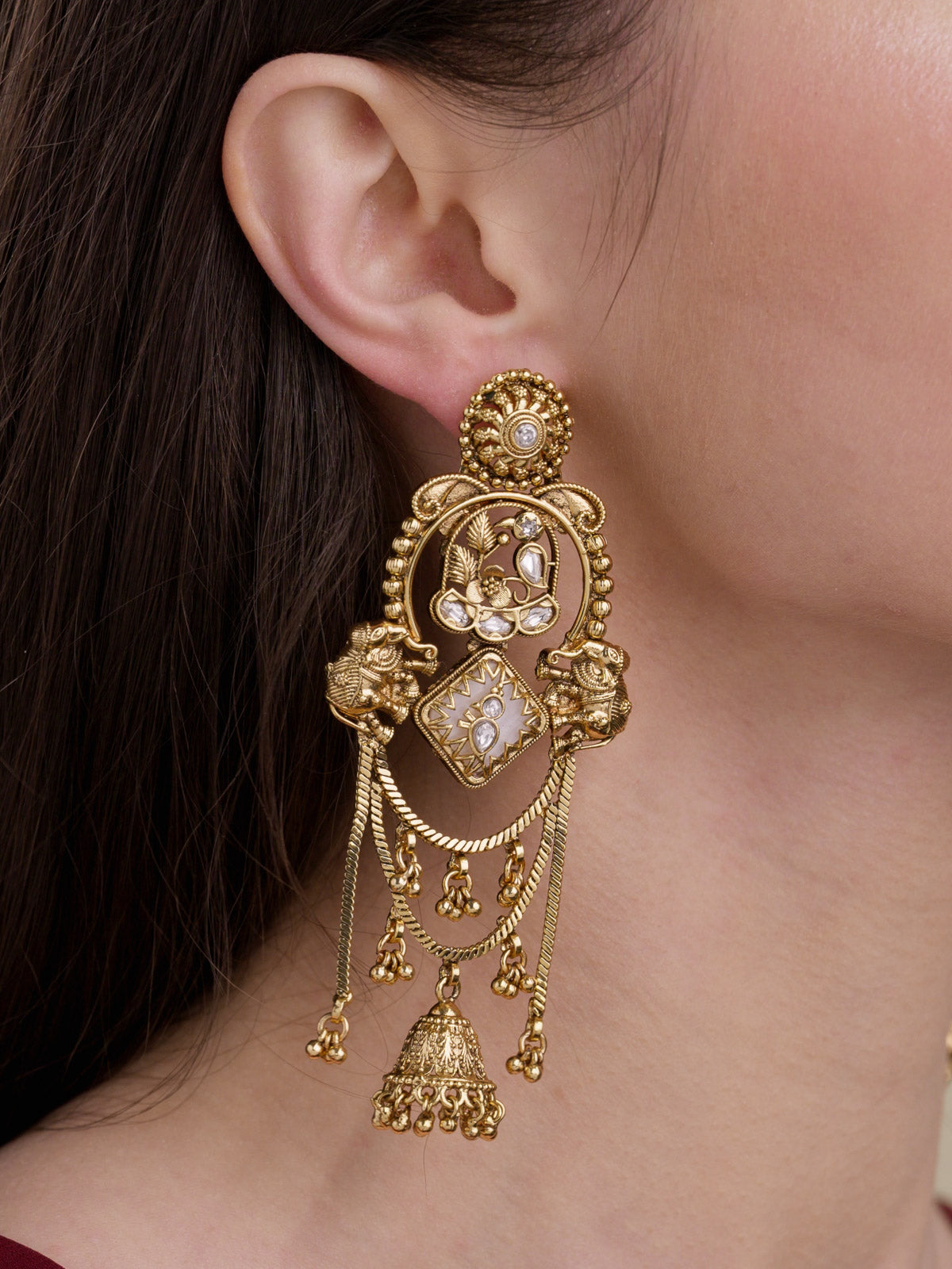 Close-up of a model wearing a matte gold tone Rajwadi statement earring with paired elephant motifs, clear stone accents, layered chain drapes and a lower jhumka drop