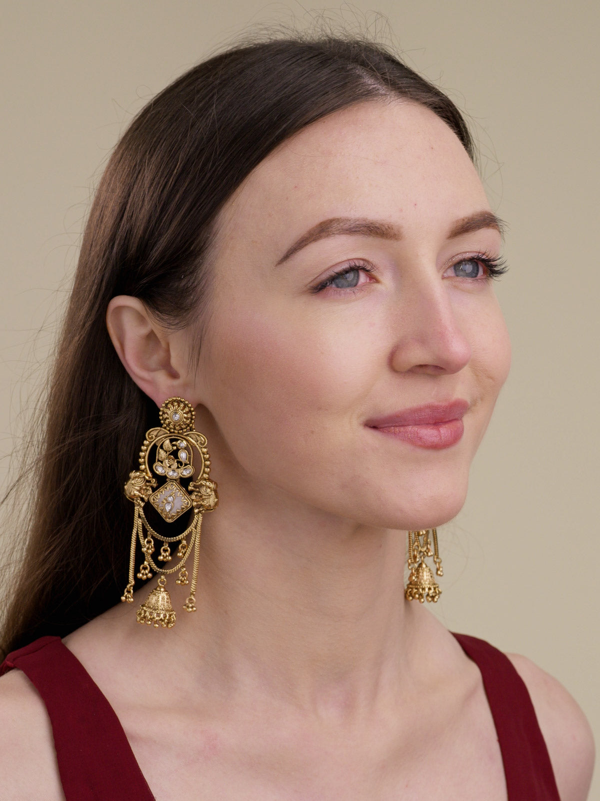 Model shot of a model wearing matte gold tone Rajwadi statement earrings with elephant motifs, clear stone accents, chain drapes and lower jhumka drops