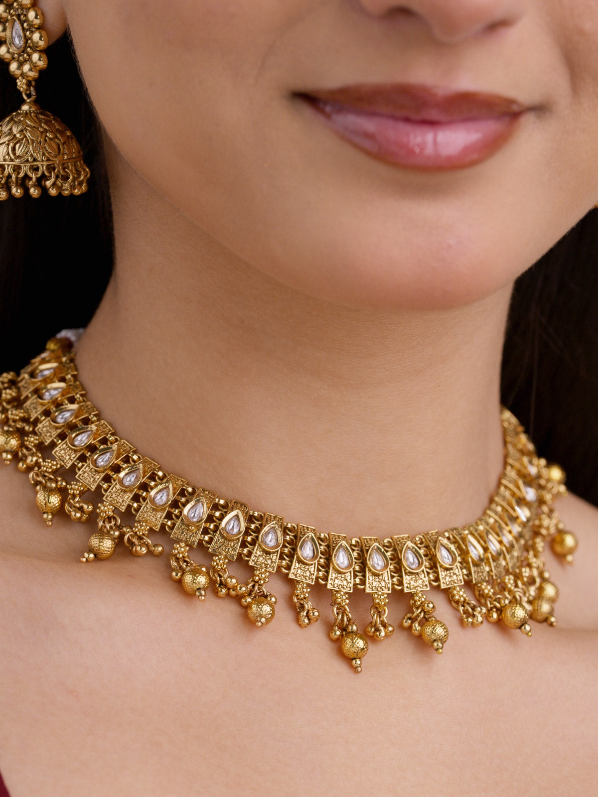 Close-up of a model wearing a gold tone Rajwadi necklace set with teardrop stone detailing, carved square links, bead drops, and a partially visible matching jhumka earring.