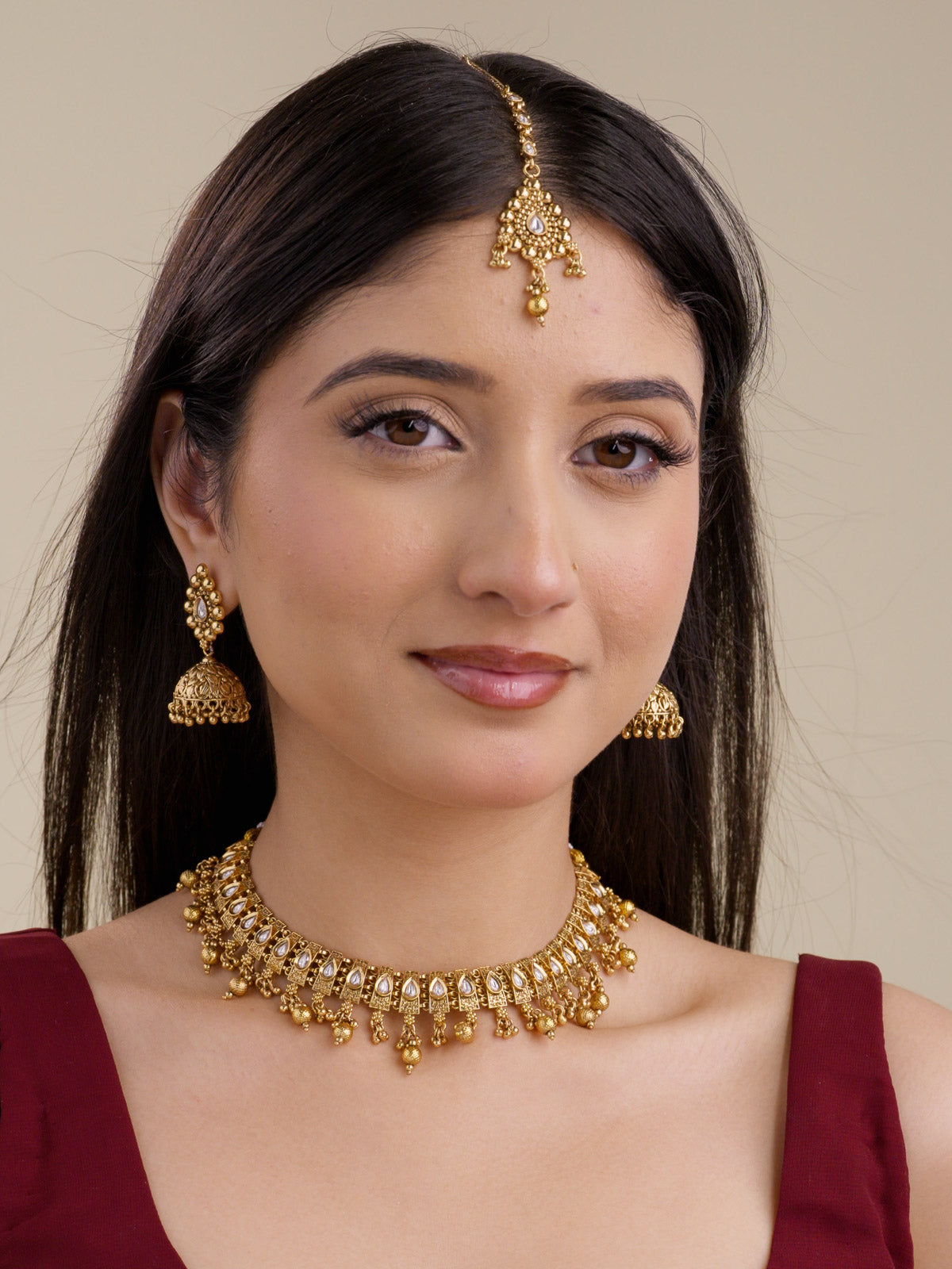 Model wearing a gold tone Rajwadi necklace set with matching jhumka earrings and maang tikka, featuring teardrop stone detailing, carved links, and bead drops.