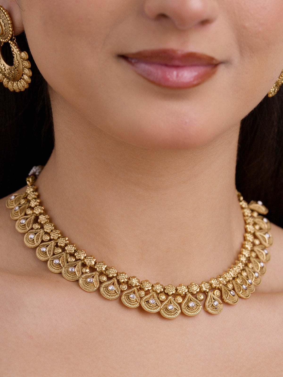 Close-up of a Rajwadi necklace set in matte gold tone, showing repeated teardrop shapes, white stone accents, floral raised detailing, and matching earrings partly visible.
