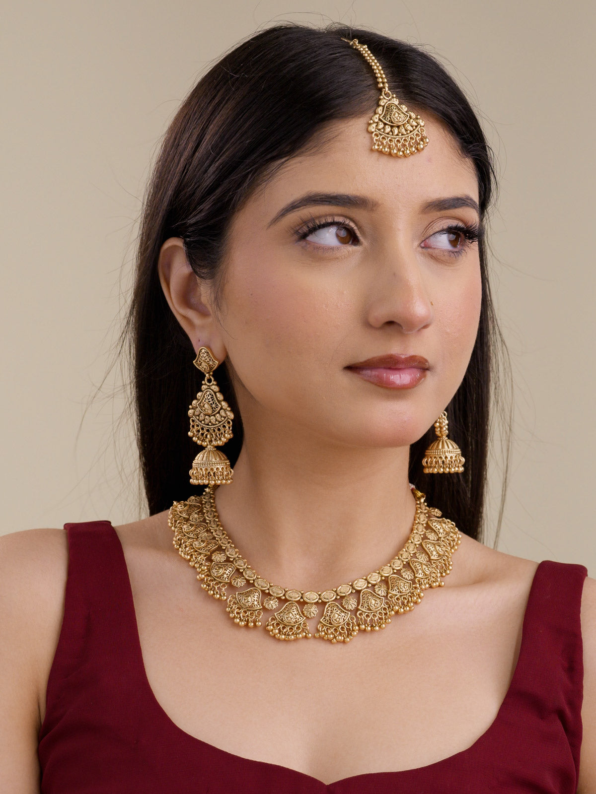 Model wearing a matte gold tone Rajwadi necklace set with teardrop-shaped filigree-style openwork detailing, small rounded metal drops, long jhumka earrings, and matching maang tikka.