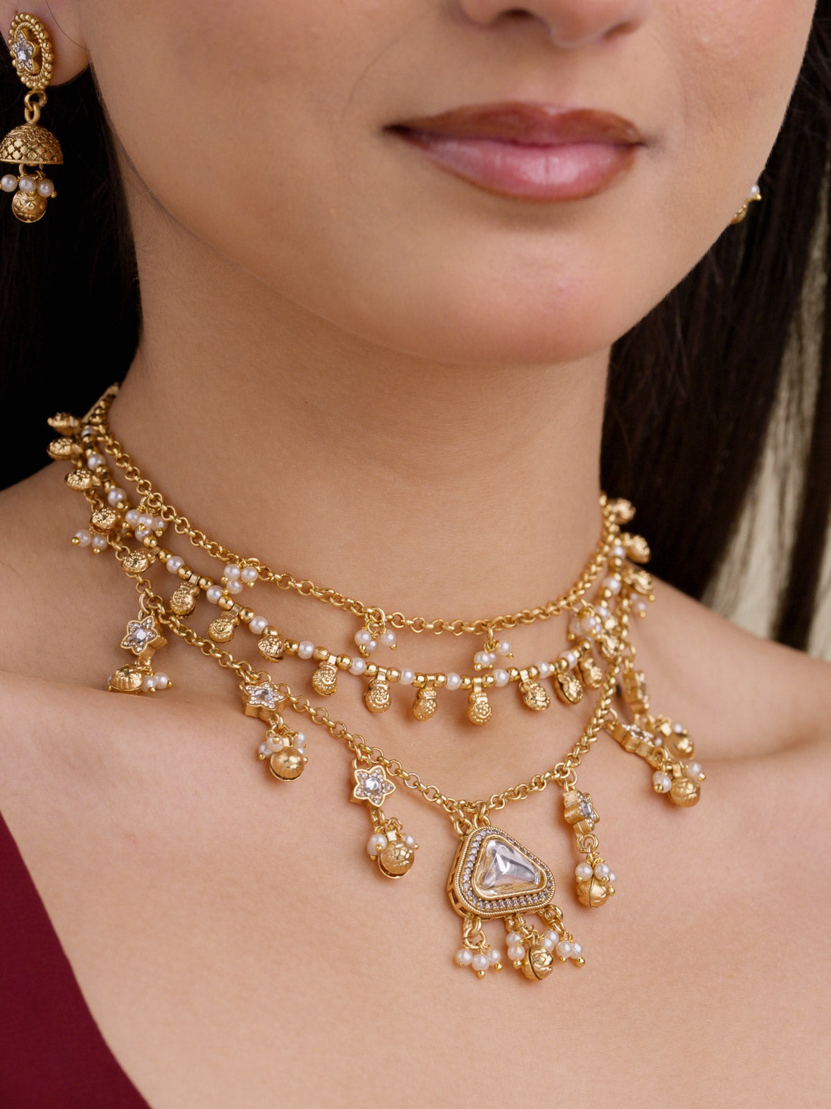 Close-up of a matte gold tone Rajwadi necklace set with layered chains, pearl-style drops, small white accent stones, a statement centre white stone, and a matching jhumka earring partly visible.