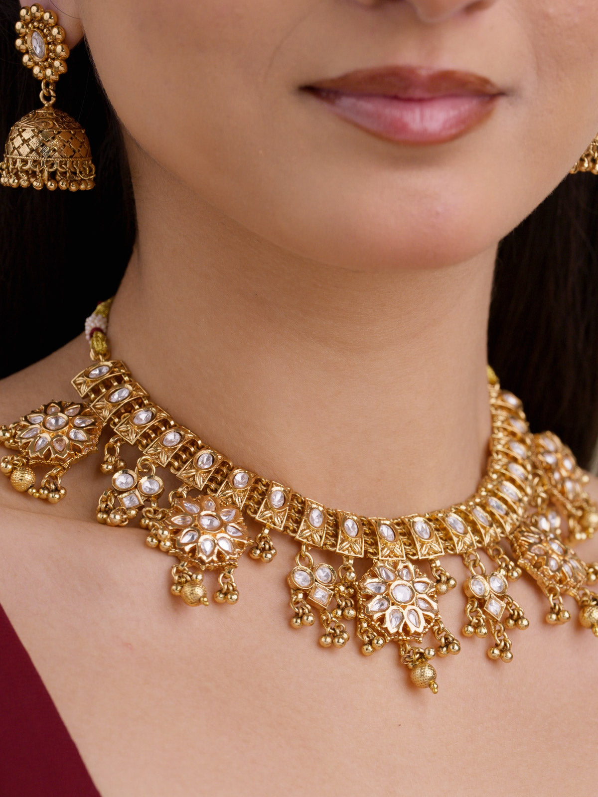 Close-up of a model wearing a gold tone Rajwadi necklace set with kundan-look stones, floral drop detailing, small gold tone metal drops, and a matching jhumka earring.