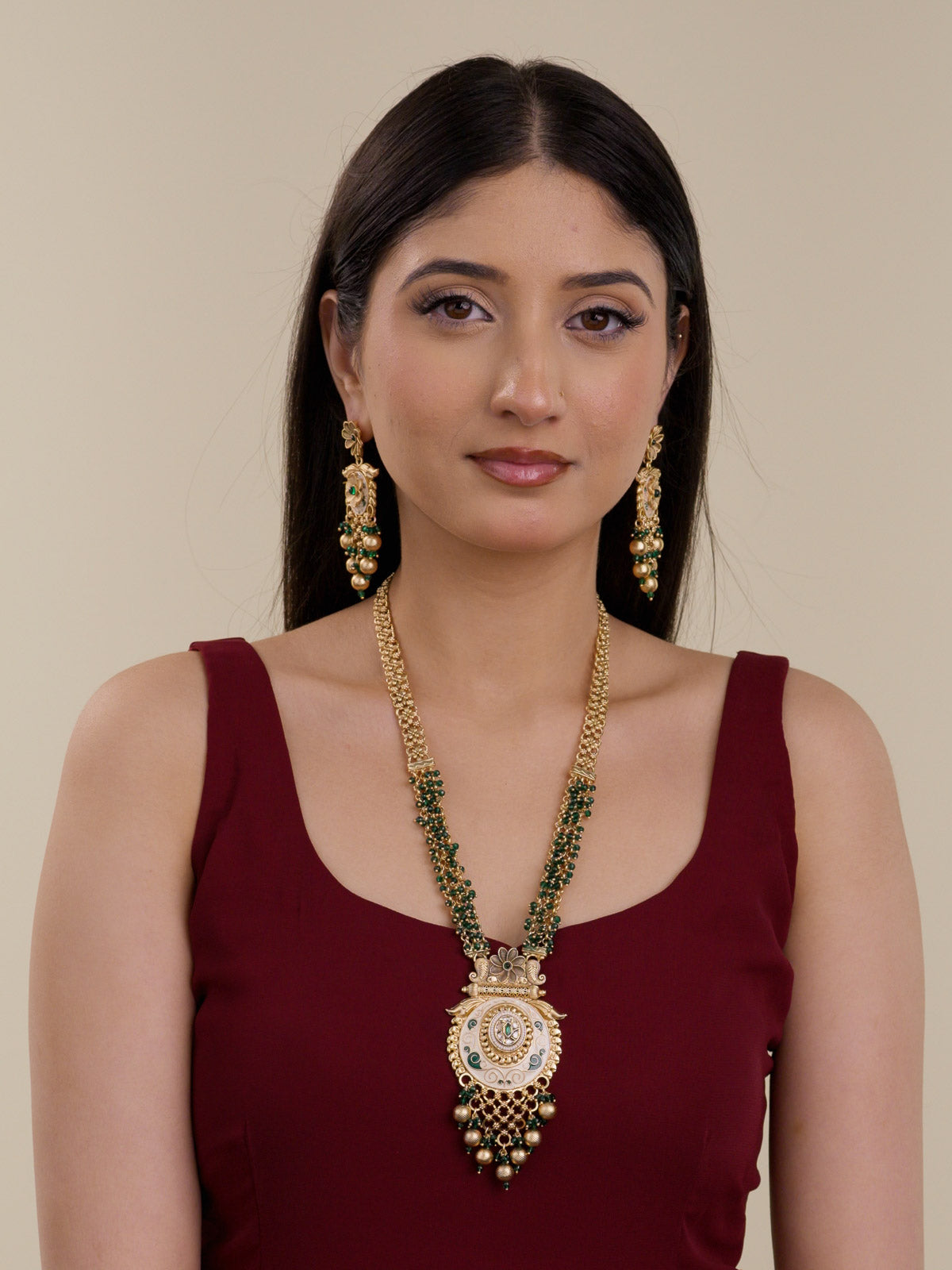 Model wearing a Rajwadi long necklace set with matte gold tone chainwork, green bead strands, Meenakari-style pendant detailing, and matching dangling earrings against a solid maroon outfit.