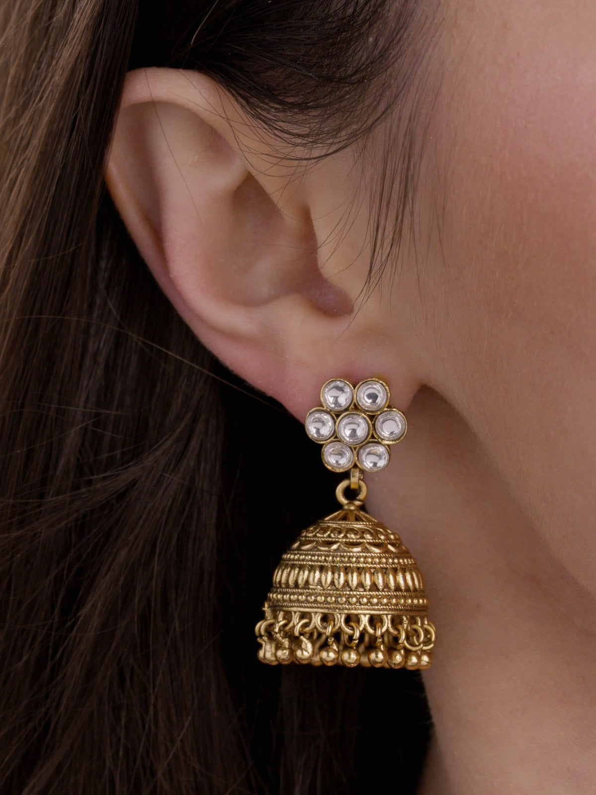 Matte gold tone Rajwadi jhumka earring with clear stone cluster stud top, carved dome detail, and small round metal drops on model close-up.