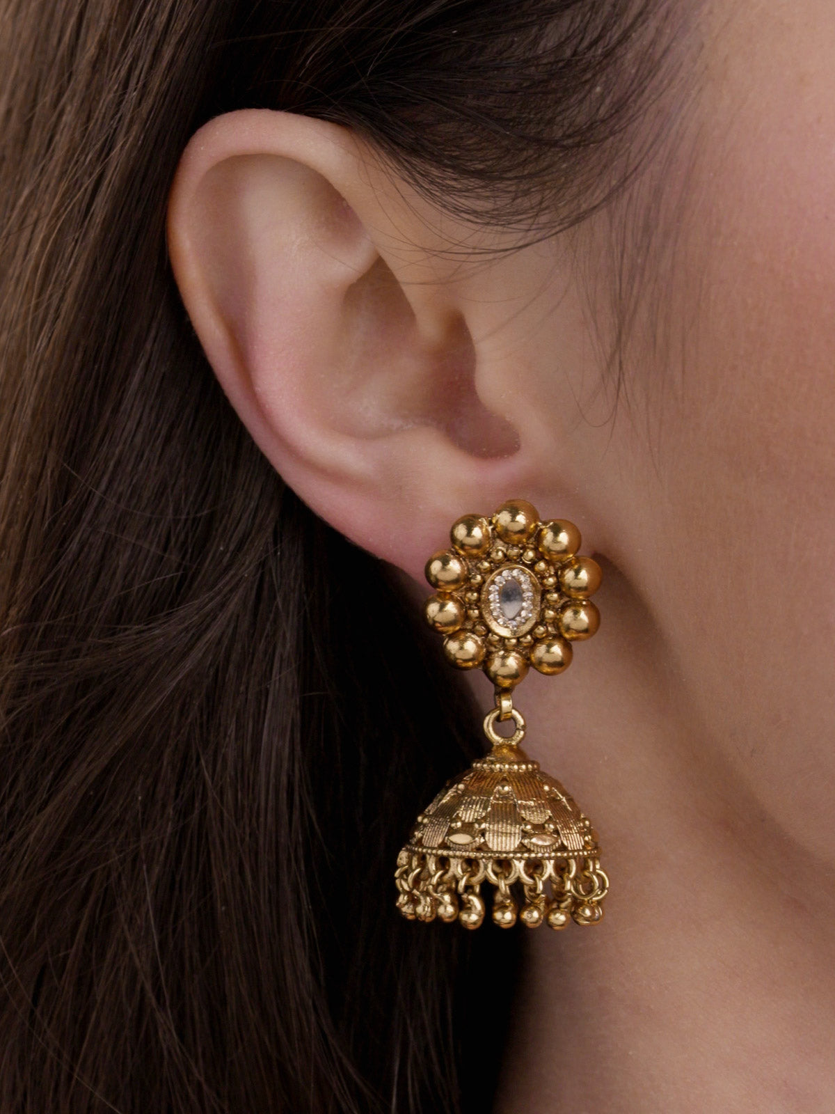 Matte gold Rajwadi jhumka earring with round metal-ball stud, clear oval stone centre, dome drop, and small round metal drops on model close-up
