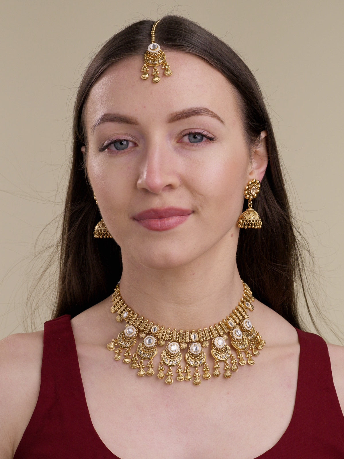 Model wearing matte gold Rajwadi necklace set with clear stone centres, crescent lower charms, shell-shaped metal drops, matching jhumka earrings, and maang tikka